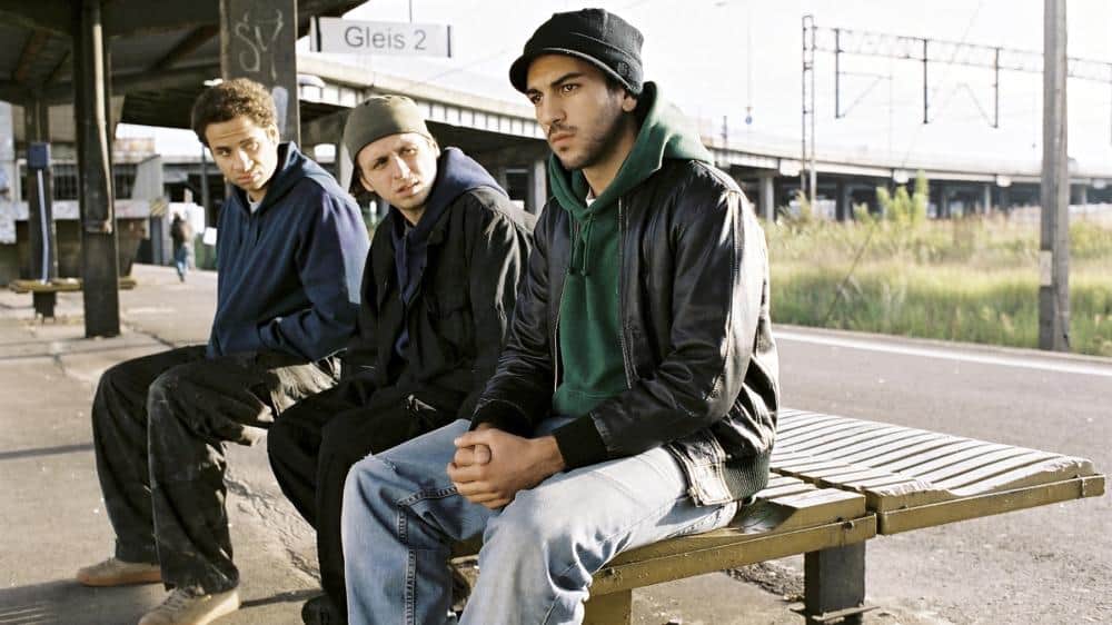 Screenshot aus dem Film Wholetrain: Die drei Hauptfiguren sitzen auf einer Bank.